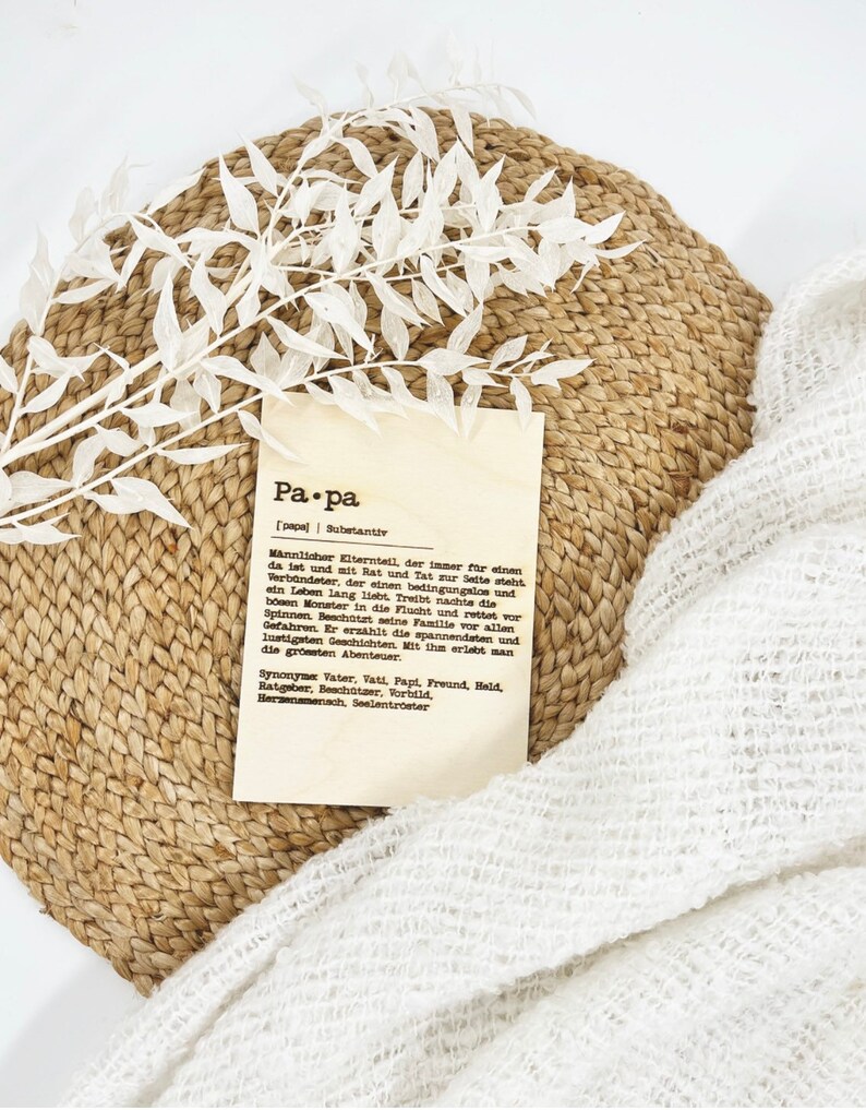 May include: A decorative arrangement featuring a woven placemat, a sprig of white leaves, a cream-colored fabric, and a card with the word "Pa.pa" and German text. The card is a definition of the word "father".