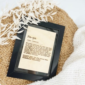 May include: A framed print with the German word "Papa" (Dad) in a black frame. The print has a definition of "Papa" in German. The frame rests on a woven basket and a white, textured blanket, with white decorative sprigs.