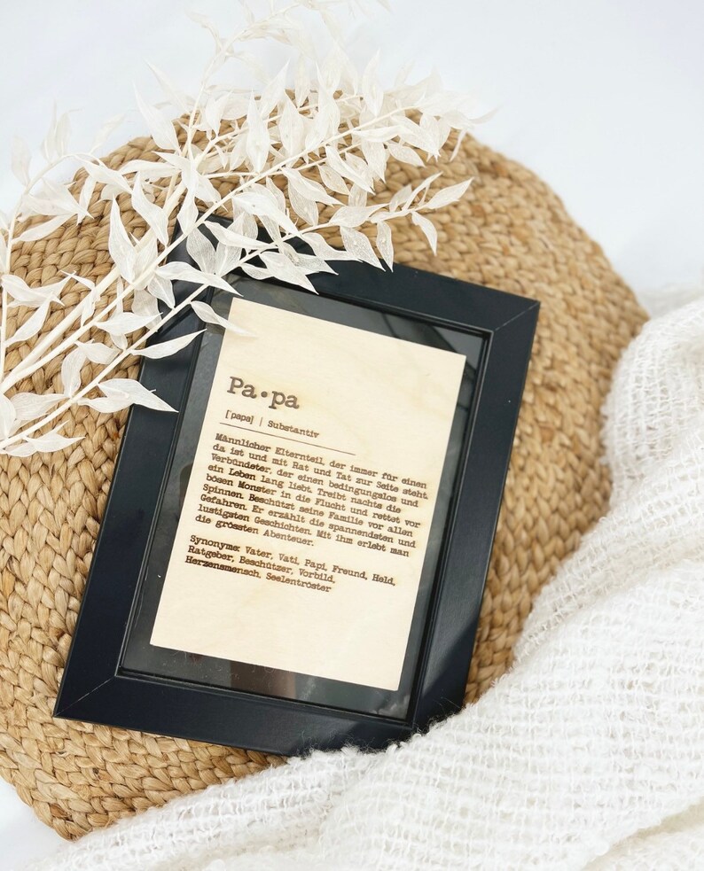 May include: A framed print with the German word "Papa" (Dad) in a black frame. The print has a definition of "Papa" in German. The frame rests on a woven basket and a white, textured blanket, with white decorative sprigs.
