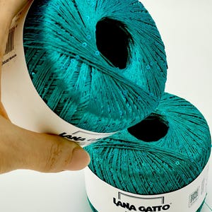May include: Two balls of teal yarn with integrated sequins, featuring a white paper label with the text Lana Gatto Paillettes Made in Italy and the number 30785. The yarn has a metallic sheen and a smooth, twisted texture throughout the skeins.