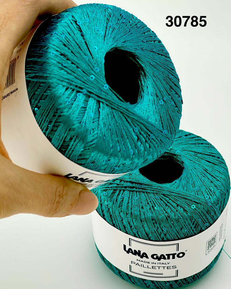 May include: Two balls of teal yarn with integrated sequins, featuring a white paper label with the text Lana Gatto Paillettes Made in Italy and the number 30785. The yarn has a metallic sheen and a smooth, twisted texture throughout the skeins.