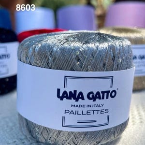 May include: A ball of silver metallic yarn with small clear sequins, wrapped in a white paper label that reads Lana Gatto Made in Italy Paillettes. The yarn has a shiny, reflective texture and is positioned in front of other yarn balls.