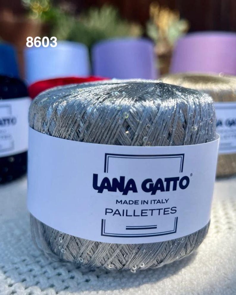 May include: A ball of silver metallic yarn with small clear sequins, wrapped in a white paper label that reads Lana Gatto Made in Italy Paillettes. The yarn has a shiny, reflective texture and is positioned in front of other yarn balls.