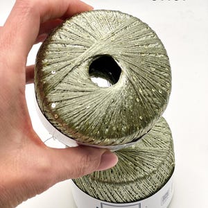 May include: Two balls of olive green yarn with integrated small sequins. The yarn features a metallic sheen and is wrapped with a white paper label that reads Lana Gatto Made in Italy Paillettes. The number 31161 is printed in the upper right corner.