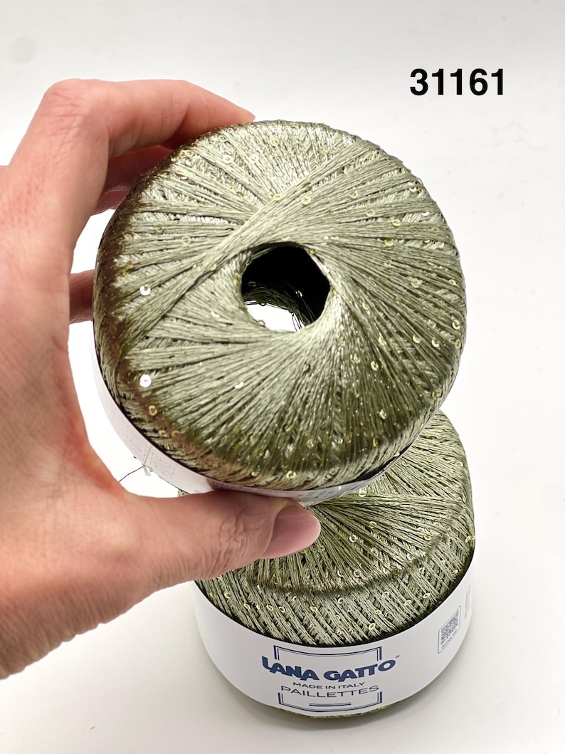 May include: Two balls of olive green yarn with integrated small sequins. The yarn features a metallic sheen and is wrapped with a white paper label that reads Lana Gatto Made in Italy Paillettes. The number 31161 is printed in the upper right corner.