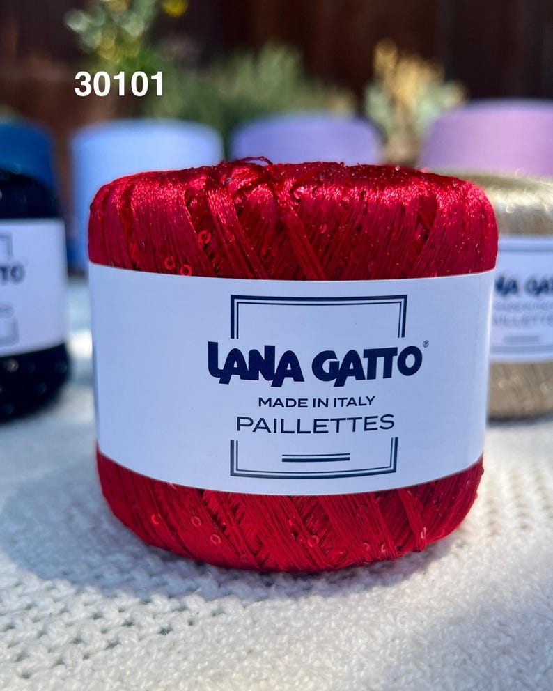 May include: A vibrant red ball of Lana Gatto Paillettes yarn featuring a white paper label with the text 30101, Lana Gatto, Made in Italy, and Paillettes. The yarn has a shiny, metallic texture with small, reflective sequins integrated throughout.
