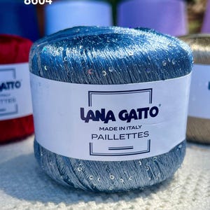 May include: A ball of metallic blue yarn with integrated iridescent sequins. The white paper label features the text LANA GATTO, MADE IN ITALY, PAILLETTES, and the number 8604. The yarn has a shiny, reflective texture and is wound into a cylindrical shape.