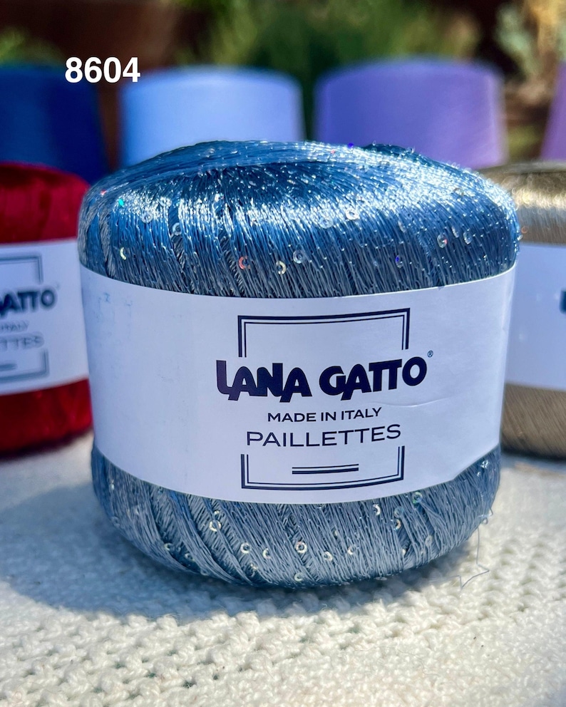 May include: A ball of metallic blue yarn with integrated iridescent sequins. The white paper label features the text LANA GATTO, MADE IN ITALY, PAILLETTES, and the number 8604. The yarn has a shiny, reflective texture and is wound into a cylindrical shape.