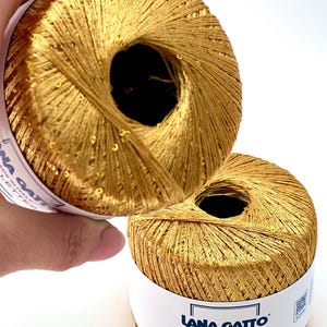 May include: Two balls of metallic gold yarn with integrated sequins. Each ball features a white paper label with the text Lana Gatto Made in Italy Paillettes and the number 30789 printed in black. The yarn has a shiny, reflective texture.