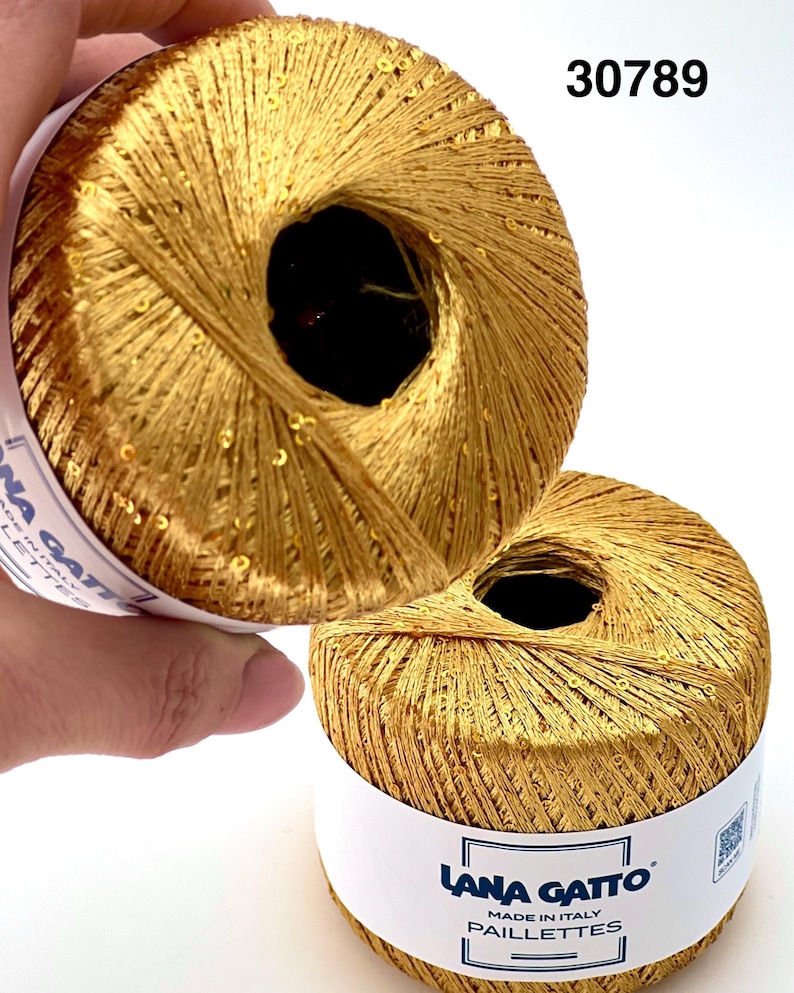 May include: Two balls of metallic gold yarn with integrated sequins. Each ball features a white paper label with the text Lana Gatto Made in Italy Paillettes and the number 30789 printed in black. The yarn has a shiny, reflective texture.