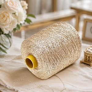 May include: A large spool of metallic gold yarn featuring a shimmering, textured finish. The yarn is wound tightly around a bright yellow cylindrical plastic core. A single, thin strand of the gold yarn extends from the spool onto the surface.