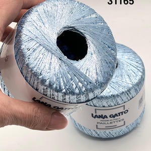 May include: Two balls of light blue metallic yarn with integrated sequins. The paper label reads Lana Gatto Made in Italy Paillettes and the number 31165. The yarn has a shiny, reflective texture and is wound into a round, compact shape.