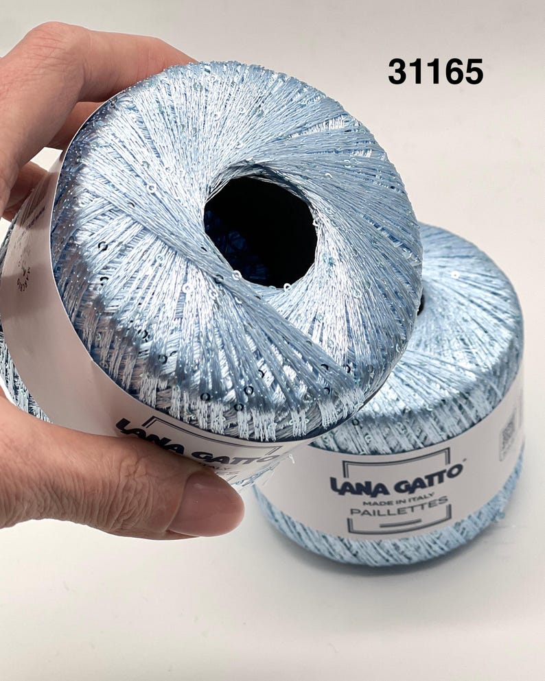 May include: Two balls of light blue metallic yarn with integrated sequins. The paper label reads Lana Gatto Made in Italy Paillettes and the number 31165. The yarn has a shiny, reflective texture and is wound into a round, compact shape.