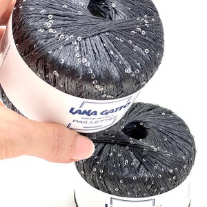 May include: Two balls of dark charcoal grey yarn featuring integrated silver sequins. Each ball has a white paper label with the text Lana Gatto Made in Italy Paillettes. The number 30103 is printed in the upper right corner of the image.