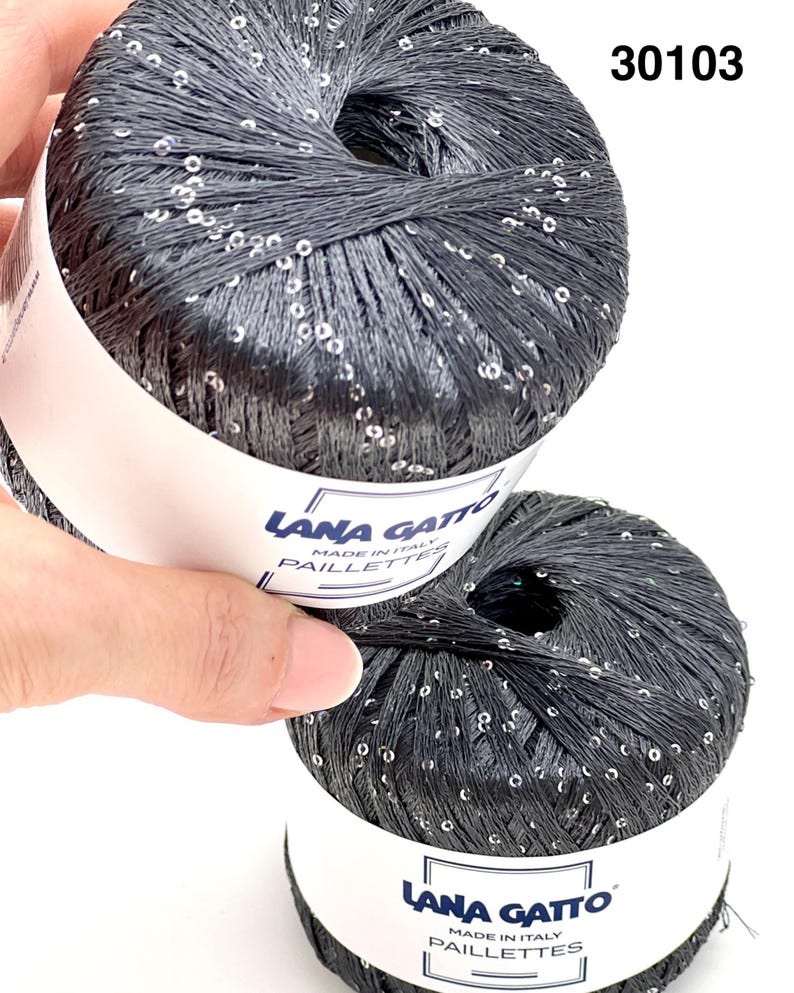 May include: Two balls of dark charcoal grey yarn featuring integrated silver sequins. Each ball has a white paper label with the text Lana Gatto Made in Italy Paillettes. The number 30103 is printed in the upper right corner of the image.