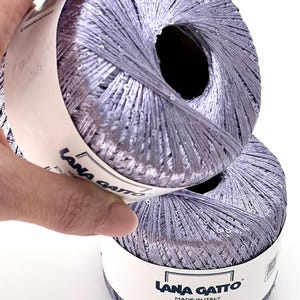 May include: Two balls of light purple yarn with a metallic sheen and embedded sequins. Each ball features a white paper label with the text Lana Gatto Paillettes Made in Italy. The number 30787 is printed in the upper right corner of the image.
