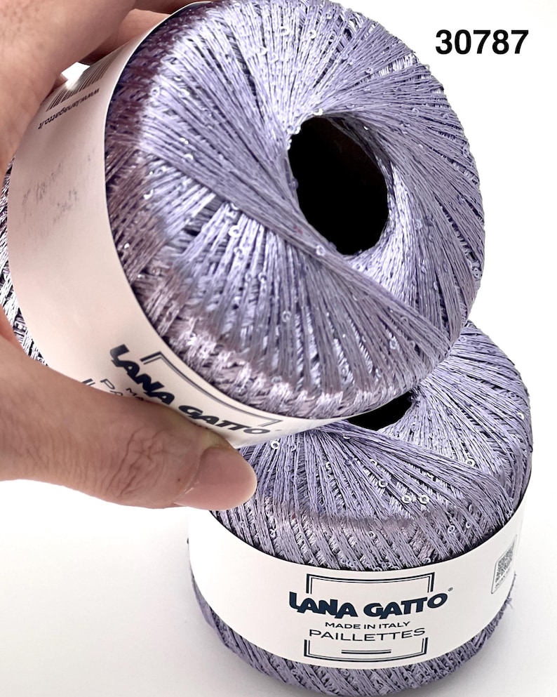 May include: Two balls of light purple yarn with a metallic sheen and embedded sequins. Each ball features a white paper label with the text Lana Gatto Paillettes Made in Italy. The number 30787 is printed in the upper right corner of the image.