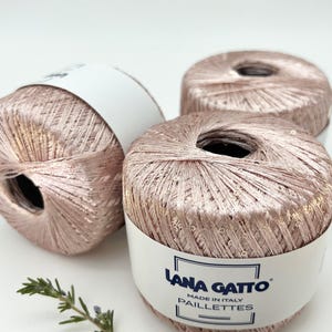May include: Three balls of metallic rose gold yarn with integrated sequins, each wrapped in a white paper band labelled Lana Gatto Made in Italy Paillettes. The yarn has a shimmering, reflective texture and a soft pink hue throughout the fibres.