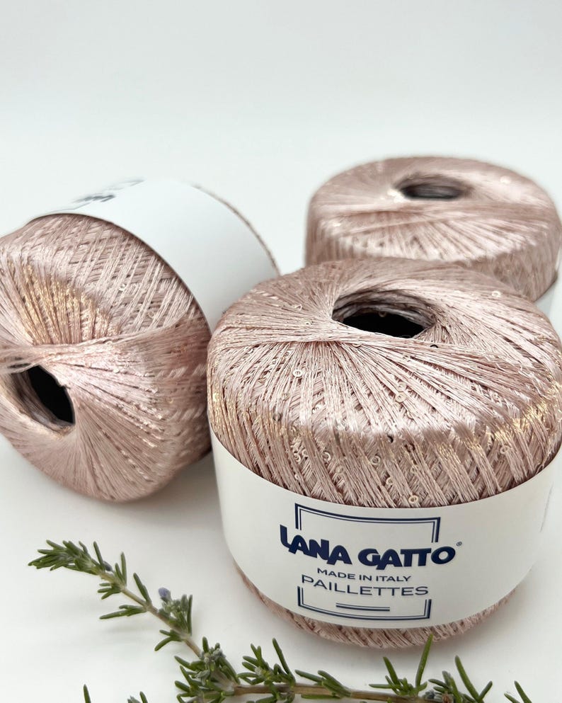 May include: Three balls of metallic rose gold yarn with integrated sequins, each wrapped in a white paper band labelled Lana Gatto Made in Italy Paillettes. The yarn has a shimmering, reflective texture and a soft pink hue throughout the fibres.