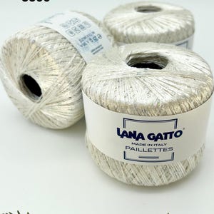 May include: Three balls of silver metallic yarn labelled Lana Gatto Paillettes, Made in Italy. Each ball features a shiny, reflective texture with subtle iridescent flecks. The label on the front ball displays the brand name and product line.