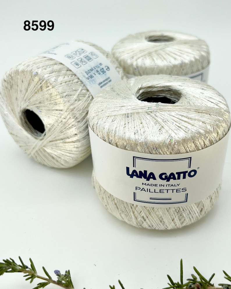 May include: Three balls of silver metallic yarn labelled Lana Gatto Paillettes, Made in Italy. Each ball features a shiny, reflective texture with subtle iridescent flecks. The label on the front ball displays the brand name and product line.