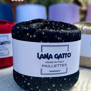 May include: A ball of black yarn with embedded silver sequins, wrapped in a white paper label featuring the text Lana Gatto, Made in Italy, and Paillettes. The yarn has a metallic sheen and a textured appearance with visible reflective accents.