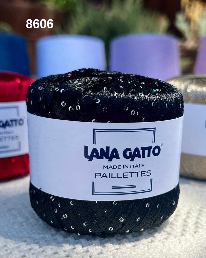 May include: A ball of black yarn with embedded silver sequins, wrapped in a white paper label featuring the text Lana Gatto, Made in Italy, and Paillettes. The yarn has a metallic sheen and a textured appearance with visible reflective accents.