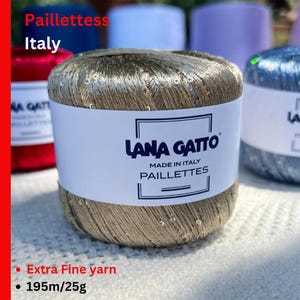May include: A ball of metallic gold Lana Gatto Paillettes yarn with small sequins. The white paper label features the text Lana Gatto, Made in Italy, and Paillettes. The yarn is labeled as extra fine with a length of 195m and a weight of 25g.
