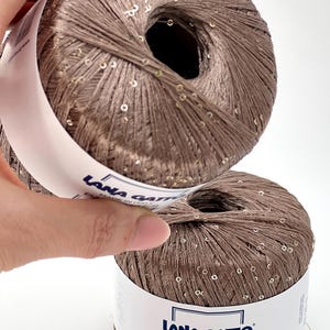 May include: Two balls of light brown yarn with integrated small gold sequins. The white paper label features the text Lana Gatto, Made in Italy, and Paillettes. The number 30100 is printed in the upper right corner of the image.