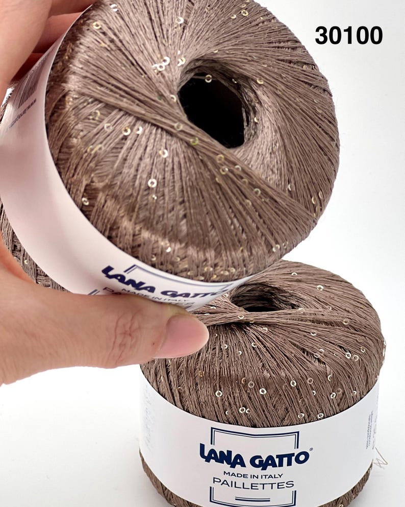 May include: Two balls of light brown yarn with integrated small gold sequins. The white paper label features the text Lana Gatto, Made in Italy, and Paillettes. The number 30100 is printed in the upper right corner of the image.