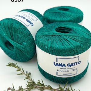 May include: Three balls of emerald green yarn with embedded sequins, each wrapped in a white paper label featuring the text Lana Gatto Paillettes Made in Italy. The number 8937 is printed in black in the upper left corner of the image.