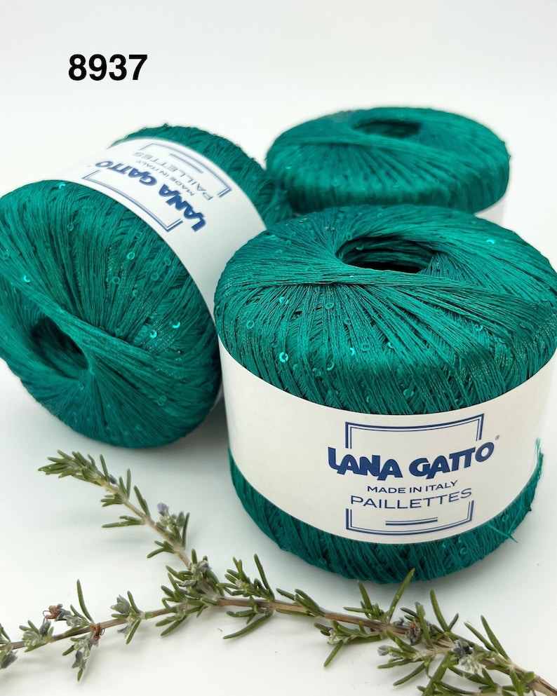 May include: Three balls of emerald green yarn with embedded sequins, each wrapped in a white paper label featuring the text Lana Gatto Paillettes Made in Italy. The number 8937 is printed in black in the upper left corner of the image.