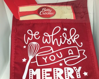 Op de afbeelding: Rode ovenwant met een wit ontwerp met de tekst "We whisk you a Merry Christmas" met een garde en sterren. De ovenwant heeft een zakje met een Betty Crocker-pakket erin.