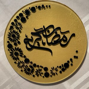 May include: A gold round plate with black Arabic calligraphy that says "Ramadan Kareem" and a black floral design around the edge.