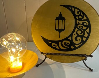 May include: A gold decorative plate with a black crescent moon and lantern design. The plate is on a black metal stand.