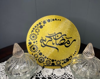 May include: A gold round wooden sign with black Arabic calligraphy that reads "Ramadan Kareem" surrounded by a black floral design. The sign is on a black stand and sitting on a white crocheted doily.