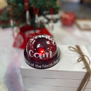 May include: A red glitter dome with the words "Santa Cam" and "Property of the North Pole" written on it. The dome has a small red light inside.