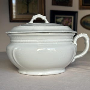 May include: A white ceramic serving dish with a lid and handle. The dish has a rounded body with a decorative handle on the side and a handle on the lid. The dish has a textured rim and a glossy finish.