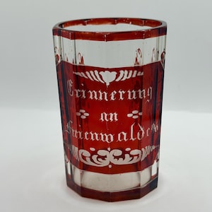 Puede incluir: Un vaso transparente y facetado con una capa roja y letras blancas. El texto dice "Erinnerung an Fmenwald". El vaso tiene un diseño decorativo en la parte superior e inferior.