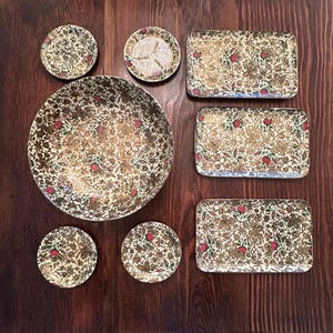 Könnte beinhalten: Ein dekoratives Servier-Set mit floralem Muster. Das Set umfasst eine große Schüssel, rechteckige Tabletts und runde Untersetzer. Das Design zeigt goldene, rote und grüne florale Elemente auf cremefarbenem Hintergrund. Die Teile sind auf einer dunklen Holzoberfläche angeordnet.