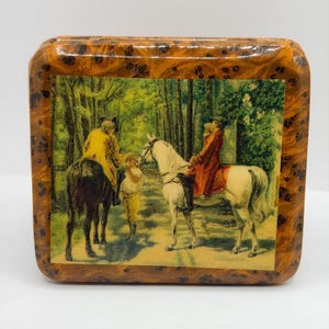 Puede incluir: Una caja cuadrada de madera con un acabado brillante de madera de raíz. La caja presenta una impresión vintage enmarcada que representa figuras a caballo en un entorno forestal. La impresión tiene un tono sepia, con acentos verdes y amarillos.
