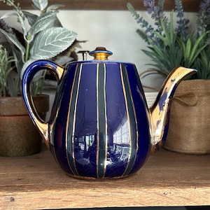 May include: A blue ceramic teapot with gold accents. The teapot has vertical gold and green stripes. The handle and spout are gold. The lid has a gold finial. The teapot is sitting on a wooden shelf.
