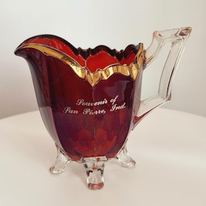 Puede incluir: Una jarra de crema de vidrio rojo rubí con asa y patas transparentes. El borde está adornado con oro. Las palabras "Souvenir of Pan Pierre, Ind." están grabadas en el lateral. Una pieza decorativa para servir.