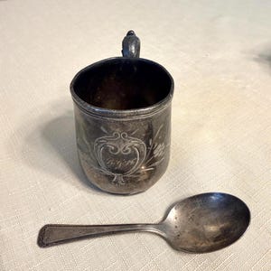 Könnte beinhalten: Eine antike Silberbecher mit Henkel und ein passender Löffel. Der Becher hat ein graviertes Design mit Initialen und floralen Akzenten. Der Löffel hat einen detaillierten Griff. Beide Artikel weisen Alters- und Gebrauchsspuren auf.