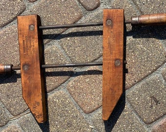 Könnte beinhalten: Eine Vintage-Holzzwinge mit Metallgewindestangen und Holzgriffen. Die Zwinge hat zwei rechteckige Holzbacken mit dem Aufdruck "Jorgensen" auf einer Seite. Die Zwinge ist für Holzbearbeitungs- und Bauprojekte konzipiert.