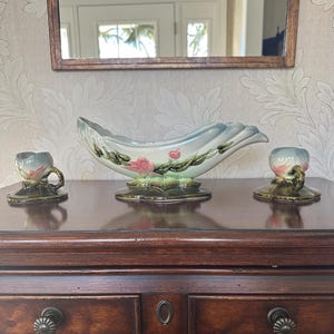 Puede incluir: Un centro de mesa de cerámica vintage con dos candelabros a juego. El centro de mesa es una fuente en forma de barco con un esmalte azul claro y blanco, decorada con flores rosas y hojas verdes. Los candelabros tienen un diseño similar.