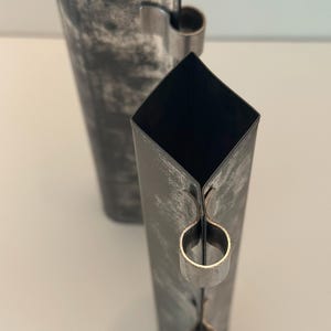 Könnte beinhalten: Zwei hohe, quadratische Metallvasen mit einem verwitterten, marmorierten Finish. Jede Vase hat einen Schlaufengriff in der Nähe der Oberseite. Die Vasen sind dunkelgrau mit hellgrauen Akzenten.