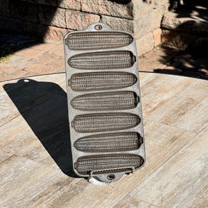 May include: A cast iron cornbread pan with seven corn cob molds. The pan is rectangular with a handle and a loop for hanging. The pan is gray and sits on a wooden table.