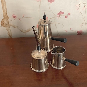 以下が含まれることがあります： 背の高いコーヒーポット、砂糖入れ、小さなクリーマーを備えた銀色のコーヒーセット。コーヒーポットと砂糖入れには黒いつまみが付いています。コーヒーポットとクリーマーのハンドルは黒色です。