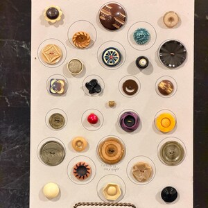 May include: A collection of vintage buttons in various shapes, sizes, and colors, arranged on a light gray background. The buttons include floral, geometric, and abstract designs, with materials like wood, plastic, and metal. Some buttons have a pearl-like finish.